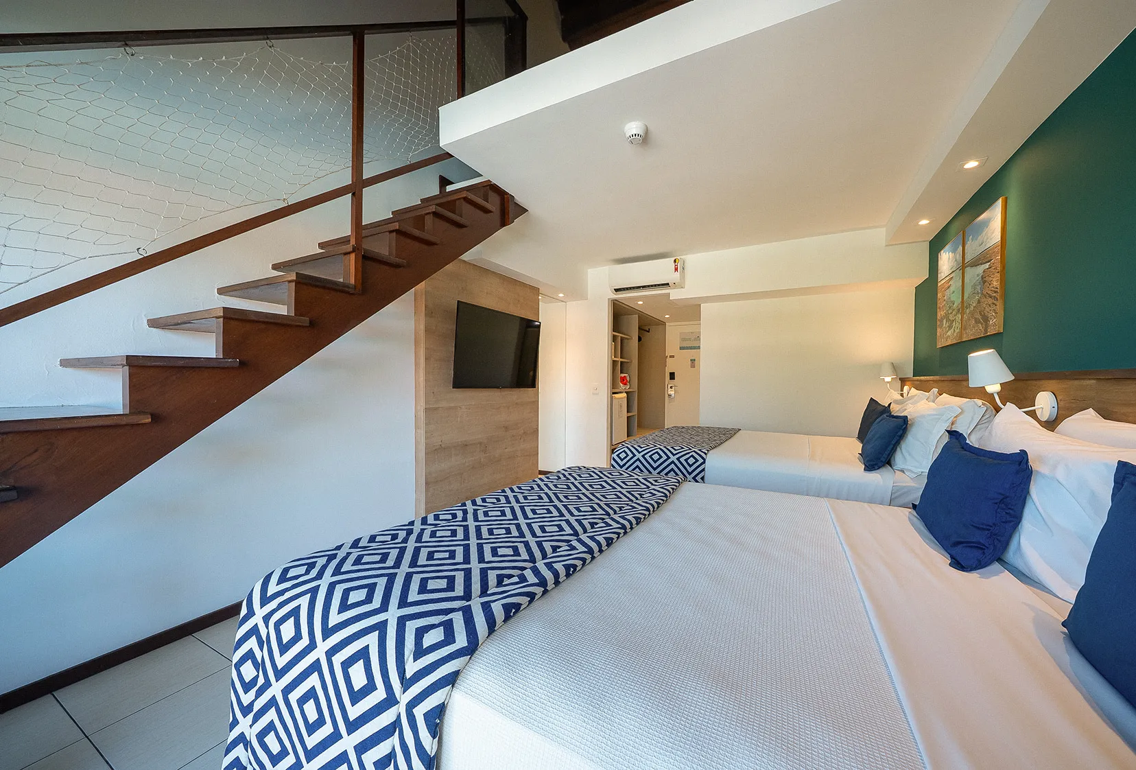 Térreo da acomodação Suíte Familiar na versão com mezanino. Duas camas queen size, duas luminárias, sendo uma para cada cama, painel de madeira com TV a cabo, espelho, roupeiro e escada levando ao primeiro andar.
