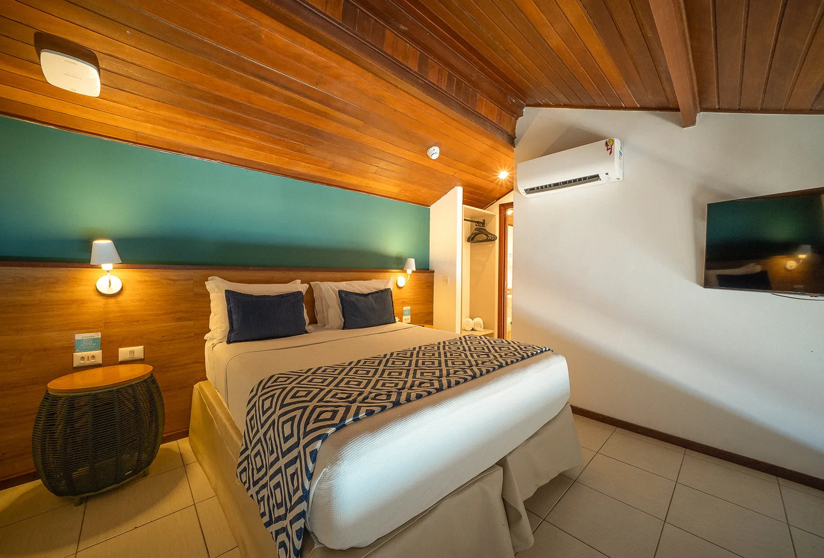 Camas de casal com duas luminárias, uma para cada lado da cama, em frente a TV a Cabo e Ar-condicionado, ao lado do roupeiro e banheiro do andar superior. O teto é mais baixo e todo de madeira.