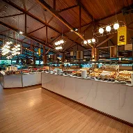 Buffet de café da manhã amplo de restaurante com balcões de mármore e diversas opções de pães e pratos quentes, sob teto de madeira e luminárias modernas.