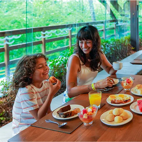 Três pessoas sorrindo em uma mesa de café da manhã no Restaurante Galés, com grandes janelas de vidro ao fundo revelando área verde. Sobre a mesa há pratos com frutas, melancia, ovos com bacon, pães de queijo, bolos, iogurte com frutas, suco de laranja e xícaras de café; um card com o número 25 aparece ao centro.