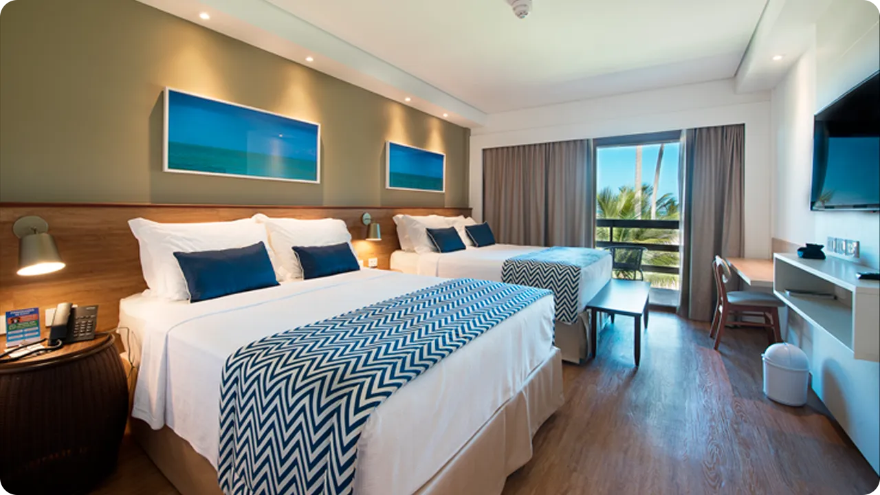 Detalhes da acomodação Praia Frente Mar, com duas camas queen size, mesa com cadeira, painel com televisão a cabo, decoração aconchegante, quadros com fotografias de praias de Maragogi e uma varanda com vista pro mar.