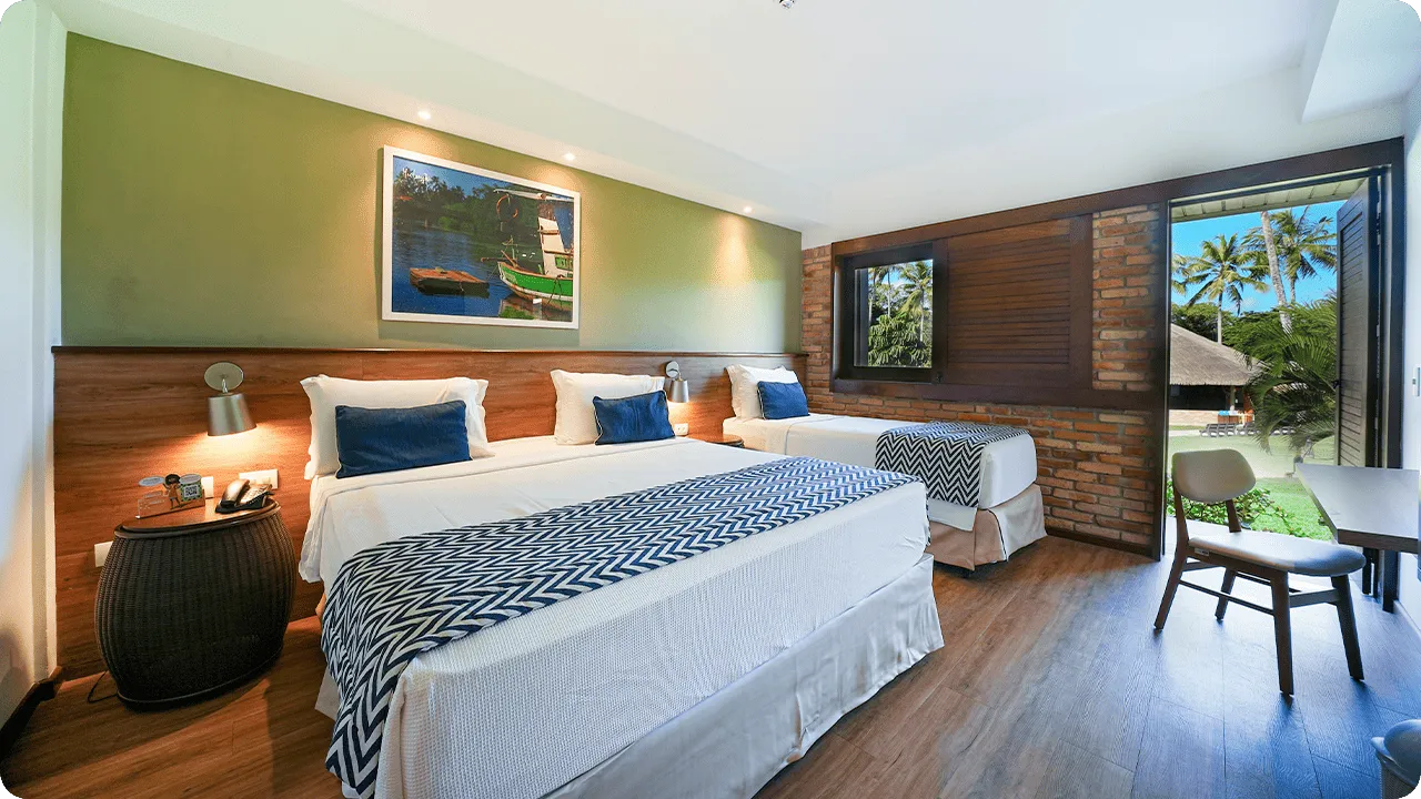 Quarto de hotel Apartamento Standard configurado para três pessoas, com cama de casal e de solteiro, decoração moderna com parede verde e de tijolinhos, e porta da varanda aberta para a área de lazer com coqueiros.