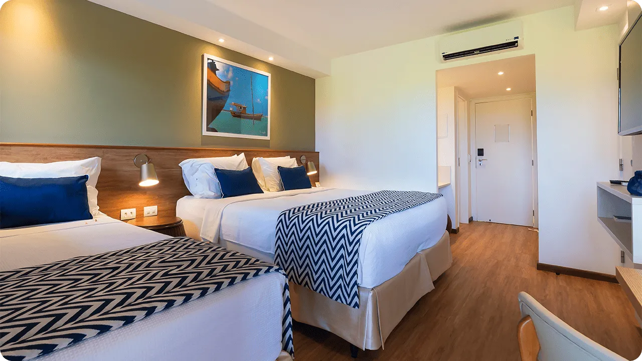 Visão interna do Apartamento Standard triplo, mostrando a cama de casal e a de solteiro, a decoração com parede verde, piso de madeira, ar-condicionado e televisão.