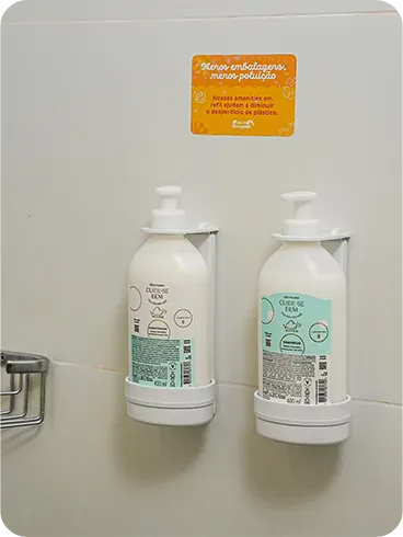 Shampoo e condicionador O Boticário em dispensers de refil na parede do banheiro, acompanhados de um aviso sobre sustentabilidade e redução de plástico.
