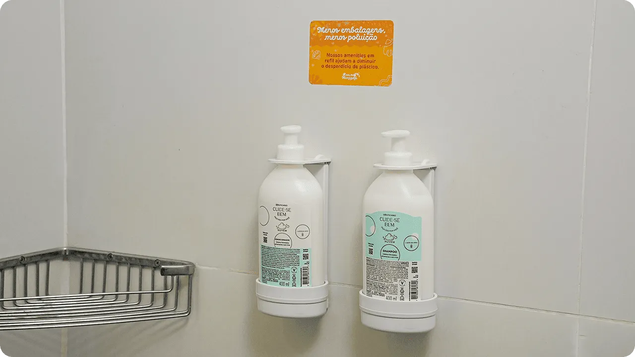 Shampoo e condicionador O Boticário em dispensers de refil na parede do banheiro, acompanhados de um aviso sobre sustentabilidade e redução de plástico.