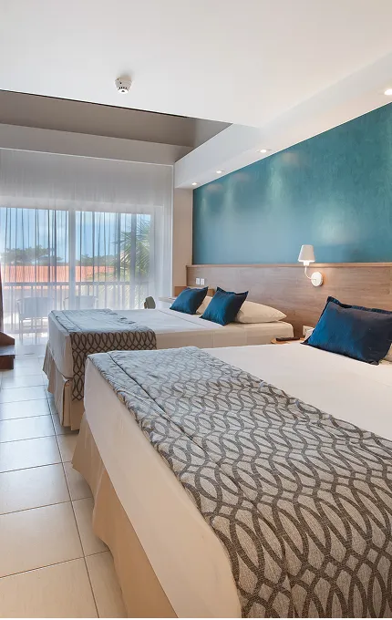 Suite Familiar, una de las habitaciones del Salinas Maceió All Inclusive Resort, con dos camas tamaño queen aconchegantes, panel de madera atrás delas, con luminária, porta da varanda y escalera para el mezanino.