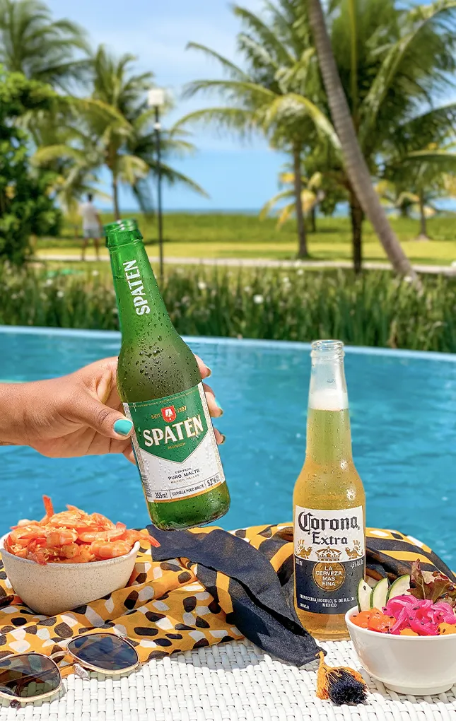 À beira de uma piscina azul, uma mão segura uma garrafa de cerveja Spaten; ao lado, uma Corona long neck, tigelas com camarão e salada sobre uma toalha estampada, óculos de sol e chapéu de palha; ao fundo, palmeiras e o mar desfocados.