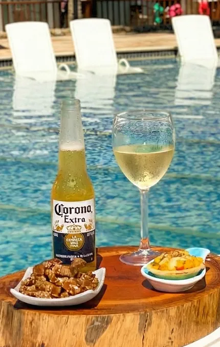 Mesa com cervejas Corona e Beck's, taça de vinho branco, caipirinha e petiscos servidos ao lado de piscina azul com espreguiçadeiras.