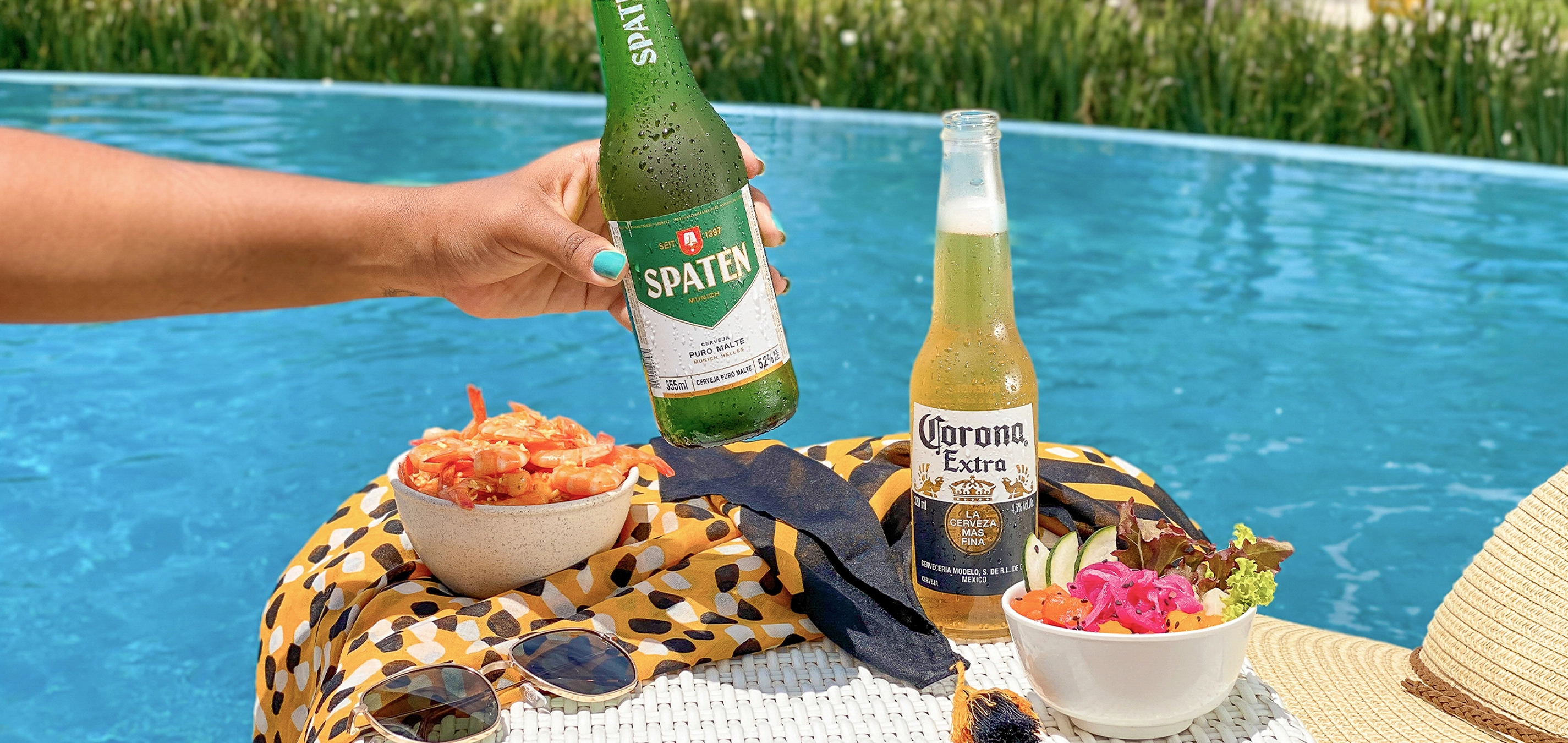 À beira de uma piscina azul, uma mão segura uma garrafa de cerveja Spaten; ao lado, uma Corona long neck, tigelas com camarão e salada sobre uma toalha estampada, óculos de sol e chapéu de palha; ao fundo, palmeiras e o mar desfocados.