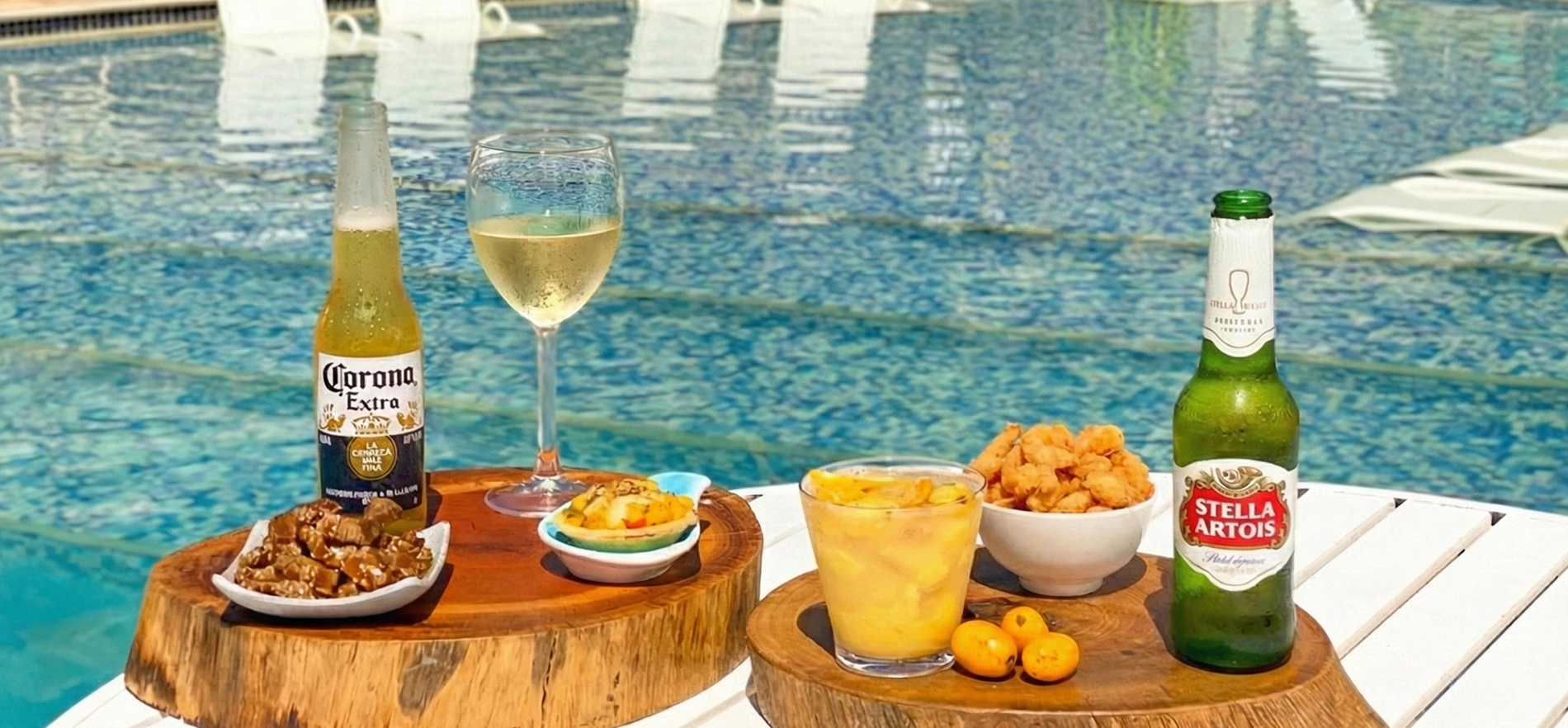 Mesa com cervejas Corona e Beck's, taça de vinho branco, caipirinha e petiscos servidos ao lado de piscina azul com espreguiçadeiras.