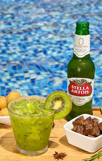 Mesa com cervejas Corona e Beck's, taça de vinho branco, caipirinha e petiscos servidos ao lado de piscina azul com espreguiçadeiras.