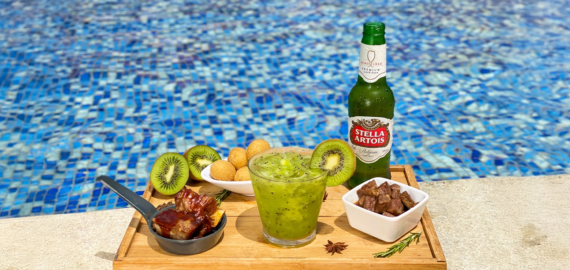 Mesa com cervejas Corona e Beck's, taça de vinho branco, caipirinha e petiscos servidos ao lado de piscina azul com espreguiçadeiras.
