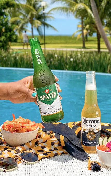 À beira de uma piscina azul, uma mão segura uma garrafa de cerveja Spaten; ao lado, uma Corona long neck, tigelas com camarão e salada sobre uma toalha estampada, óculos de sol e chapéu de palha; ao fundo, palmeiras e o mar desfocados.