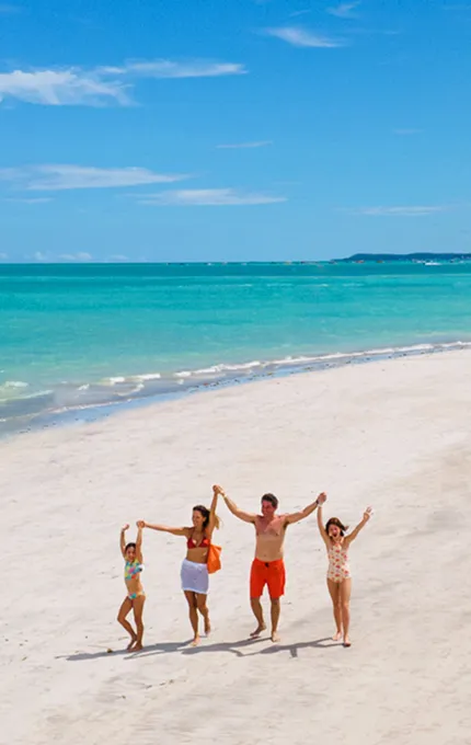 Família caminhando de mãos dadas na areia branca da praia de Maragogi com mar azul ao fundo