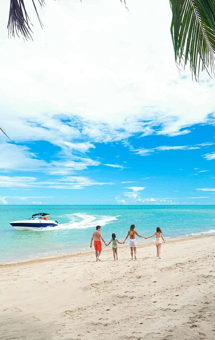 Família caminhando pela praia de Maragogi com vista para o mar e um barco ao fundo.
