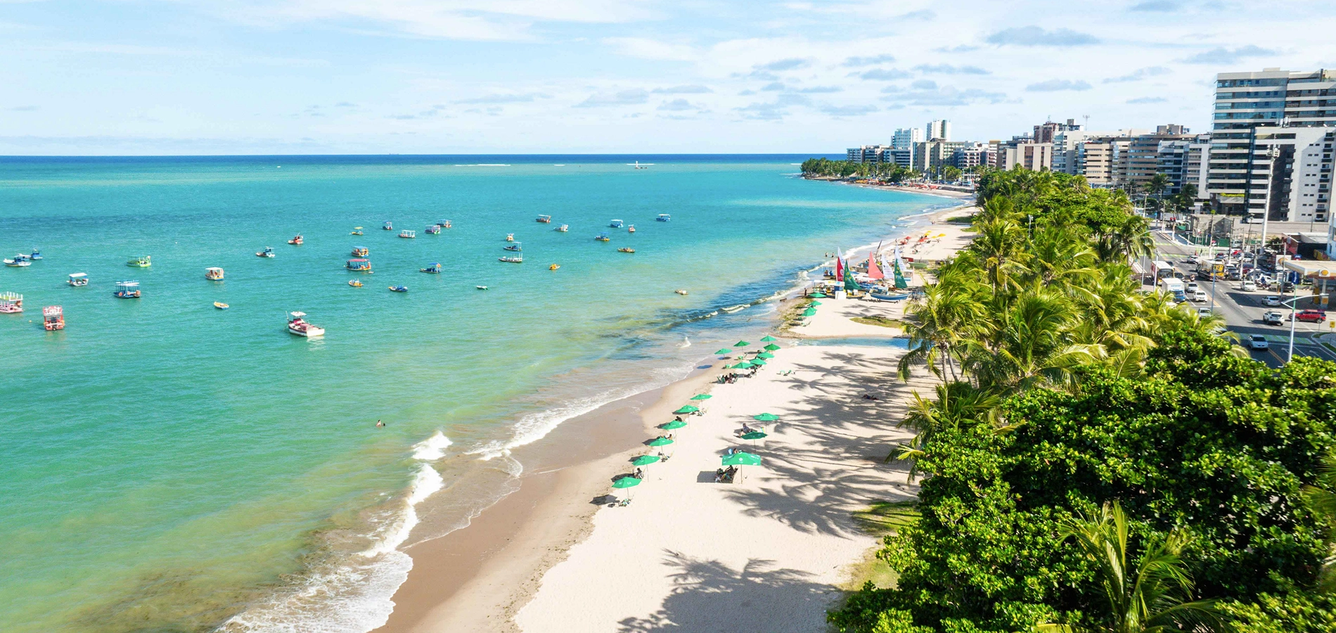 Vista aérea da orla urbana da Praia de Pajuçara, em Maceió, com o mar de cor verde-esmeralda repleto de barcos, a faixa de areia com coqueiros, e a avenida e os prédios da cidade ao fundo.