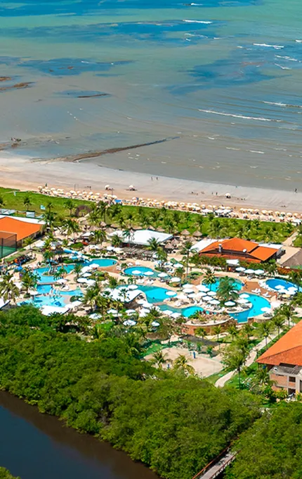  Imagem aérea do Salinas Maragogi mostrando o complexo de piscinas, dos quartos, pontos de consumo e ao fundo a praia. Área verde cercando todo o resort e o rio na parte inferior da imagem, é o rio que corta o resort. 