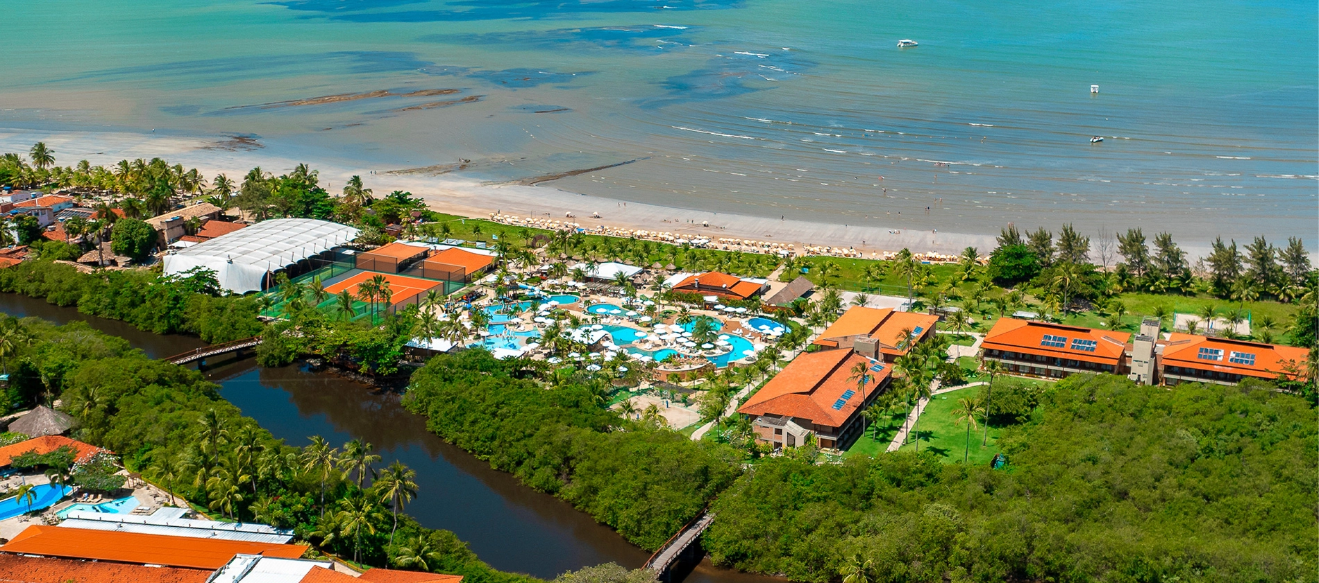  Imagem aérea do Salinas Maragogi mostrando o complexo de piscinas, dos quartos, pontos de consumo e ao fundo a praia. Área verde cercando todo o resort e o rio na parte inferior da imagem, é o rio que corta o resort. 