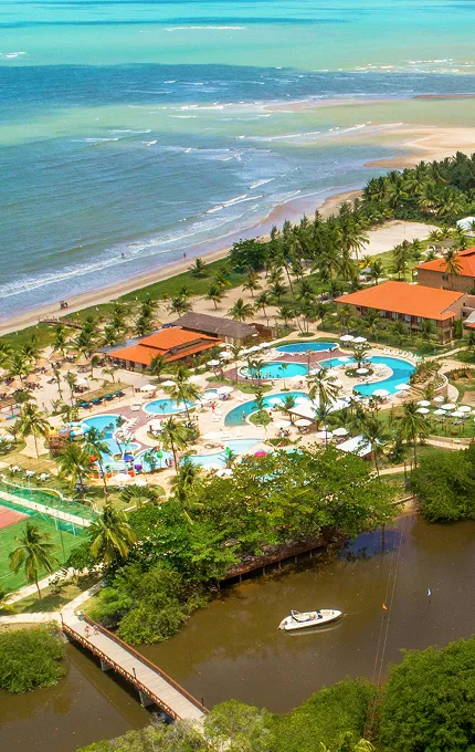 Vista aérea do complexo do resort Salinas Maragogi, que é cortado pelo Rio Maragogi e fica em frente ao mar. A imagem exibe os prédios, o grande parque aquático, as pontes e a vegetação exuberante que cerca toda a propriedade.