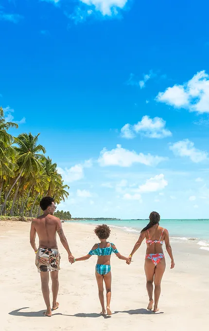 Família de costas caminhando sobre a areia branca à beira-mar da Praia de Xaréu, um dos destinos próximos ao Salinas Maragogi, em dia ensolarado.