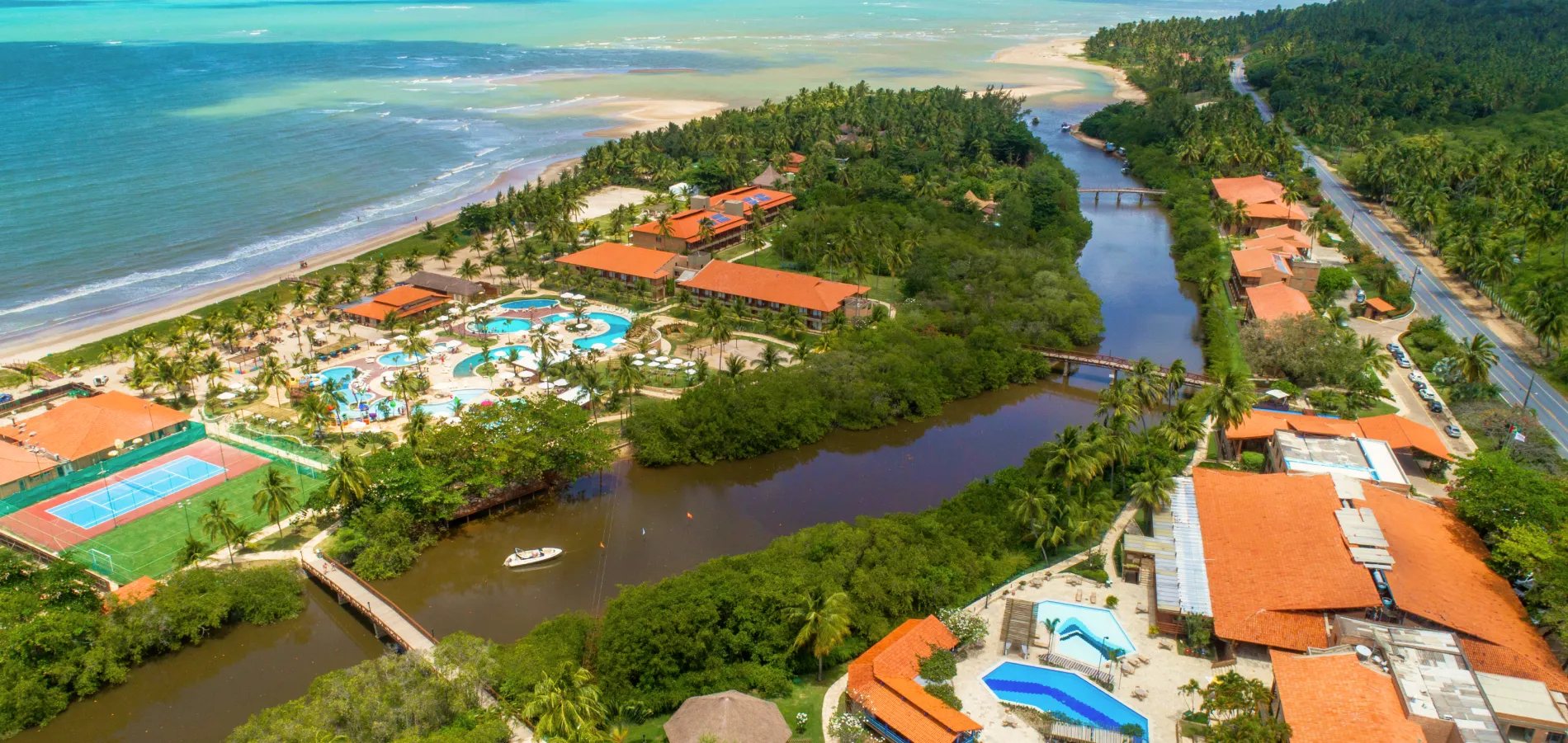 Vista aérea do complexo do resort Salinas Maragogi, que é cortado pelo Rio Maragogi e fica em frente ao mar. A imagem exibe os prédios, o grande parque aquático, as pontes e a vegetação exuberante que cerca toda a propriedade.