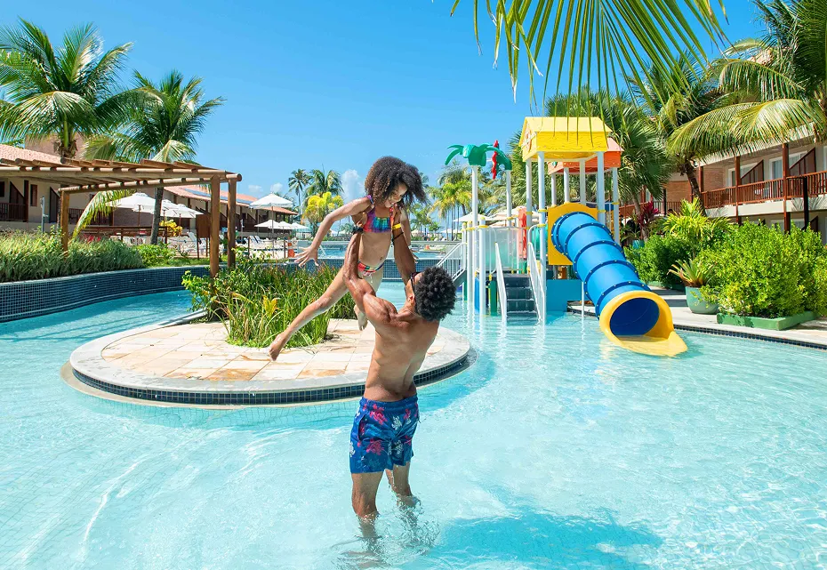 Pai e filha se divertem na piscina infantil do Salinas Maceió, com o pai levantando a criança no ar. Ao fundo, um toboágua colorido e coqueiros completam a cena de férias em família.