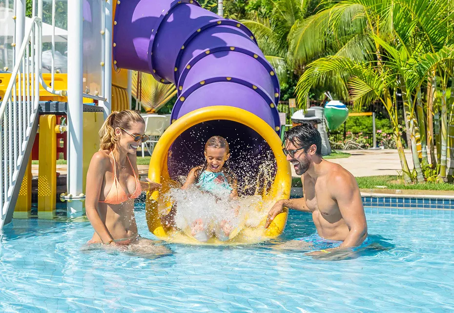 Criança saindo de tobogã colorido e sendo recebida por dois adultos sorridentes em piscina de parque aquático, com vegetação tropical ao fundo e céu azul.