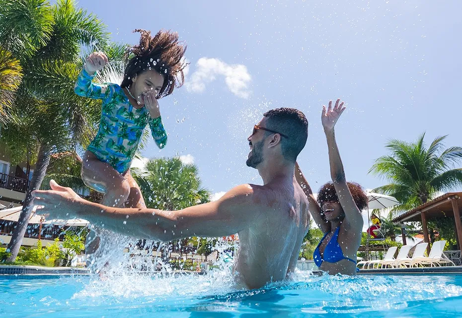 Pai e filha se divertem na piscina infantil do Salinas Maceió, com o pai levantando a criança no ar com a mãe ao lado. Ao fundo, área externa e coqueiros completam a cena de férias em família.