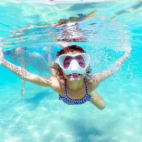 Menina sorrindo com máscara de snorkel em um mergulho nas águas cristalinas e azul-turquesa das piscinas naturais de Maceió, em uma foto que mostra tanto o fundo do mar quanto o céu azul na superfície.