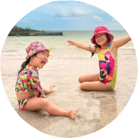 Duas crianças sorridentes sentadas na areia da praia, usando trajes de banho coloridos e chapéus, com mar calmo e céu azul ao fundo.