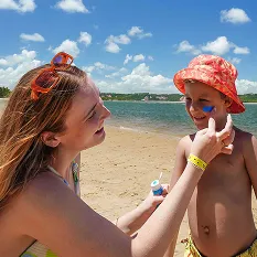 Mãe aplicando protetor solar no rosto do filho na praia do Salinas Maceió. Ambos estão sorrindo em um dia ensolarado, com o mar calmo e o céu azul ao fundo.