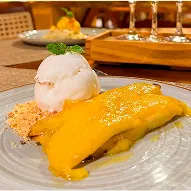 Prato fundo com arroz, molho cremoso amarelo, legumes grelhados e farofa; mão segura um garfo. Ao fundo, taça de vinho branco e salão de restaurante desfocados.