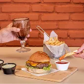 Dois hambúrgueres artesanais servidos com batatas fritas, molho e milkshake em mesa de tijolos à vista