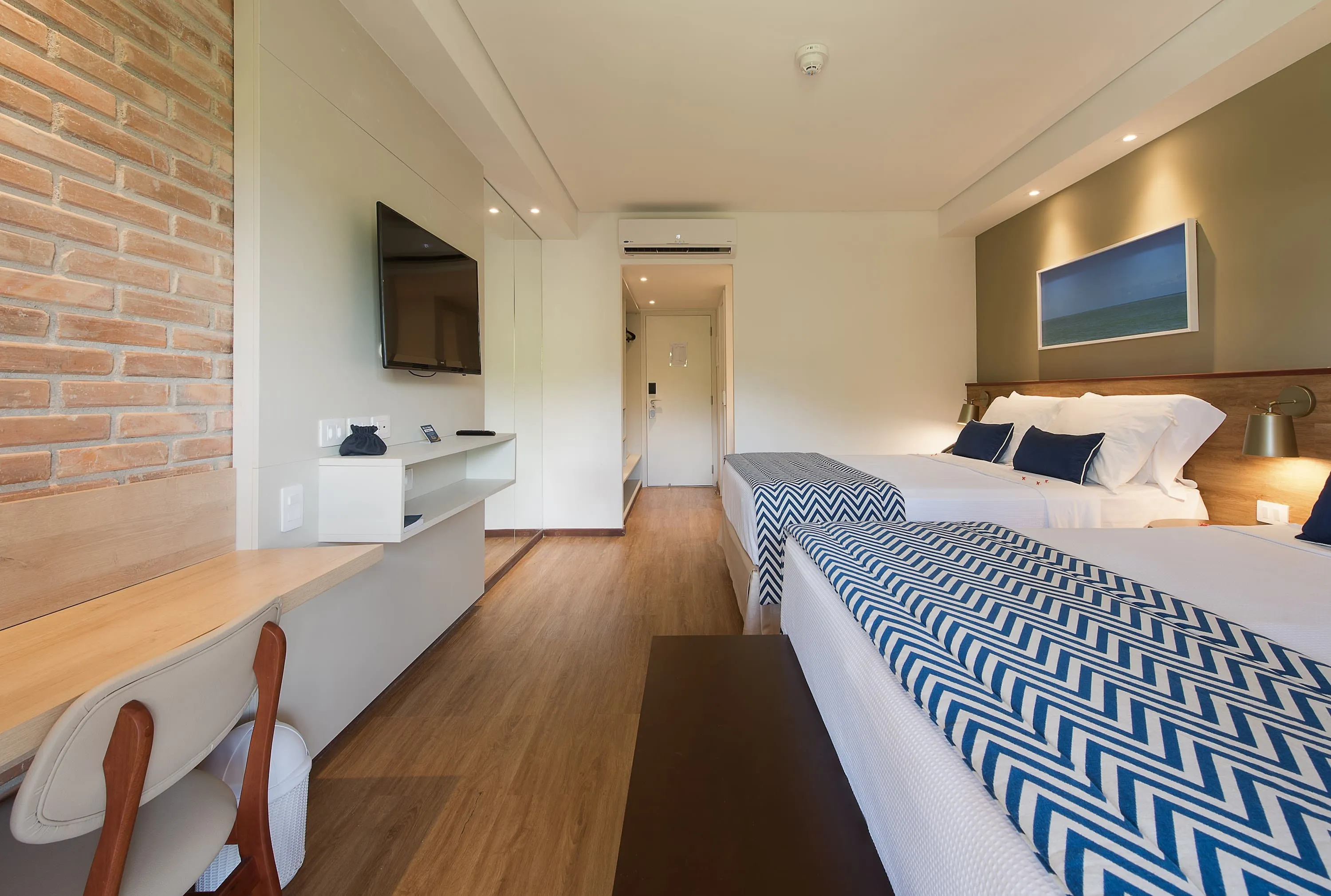 Interior del alojamiento Playa Jardín, con dos camas queen, decorado con imágenes de las playas de Maragogi y paredes de ladrillo, además de una mesa con sillas, espejos, armario y puerta de entrada.