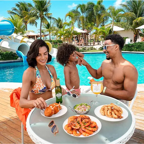 Família à beira da piscina do resort saboreando petiscos e bebidas; ao fundo toboáguas azuis, palmeiras e guarda-sóis em clima tropical