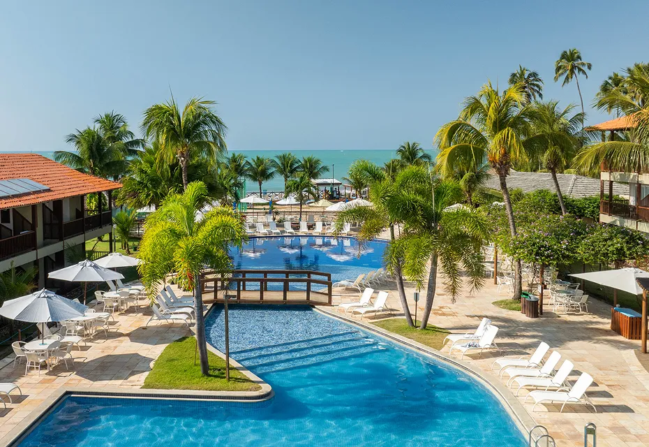 Vista aérea do resort Salinas Maceió, mostrando os prédios dos apartamentos, a ampla área de lazer com piscinas, e os coqueiros que cercam a propriedade, com o mar de Maceió ao fundo sob um céu limpo.