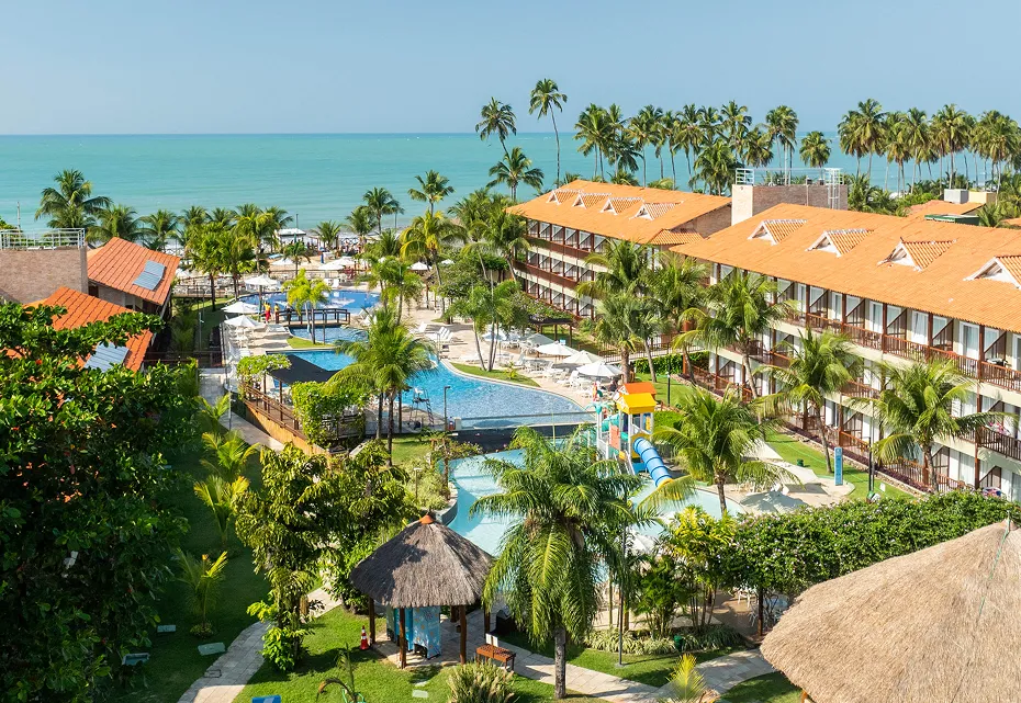 Vista aérea do resort Salinas Maceió, mostrando os prédios dos apartamentos, a ampla área de lazer com piscinas, e os coqueiros que cercam a propriedade, com o mar de Maceió ao fundo sob um céu limpo.