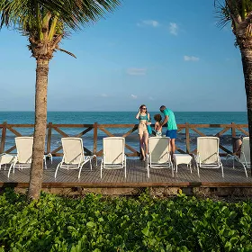 Deck de madeira no resort Salinas Maceió com espreguiçadeiras brancas voltadas para o oceano. A cena é emoldurada por dois coqueiros e uma família está no deck apreciando a vista do mar em um dia de sol.