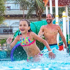 Família se divertindo no parque aquático do Salinas Maceió. Em primeiro plano, uma menina corre sorrindo na água, enquanto seus pais a seguem, rindo, em um momento de pura alegria e descontração.