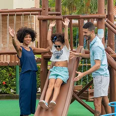 Família divertindo em parquinho infantil, a filha está descendo o escorregador com pai e mãe ao lado. Ao fundo a estrutura do resort Salinas Maceió.