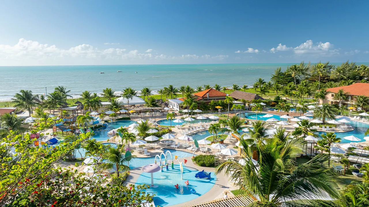 Vista aérea do complexo de piscinas do Salinas Maragogi, com espreguiçadeiras, guarda-sóis e brinquedos infantis. Há coqueiros por toda parte e um gramado ao fundo. É possível ver a praia ao fundo com sua imensidão e o céu sem nuvens.