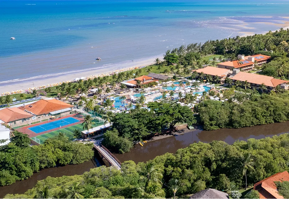 Vista aérea do Salinas Maragogi com praia de águas turquesa ao fundo, rio cercado por vegetação tropical, piscinas, quadras e estrutura integrada à natureza.