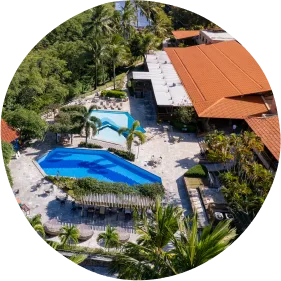 Vista aérea de resort tropical com piscinas, prédios de telhado vermelho, vegetação abundante e canal de água ao lado.