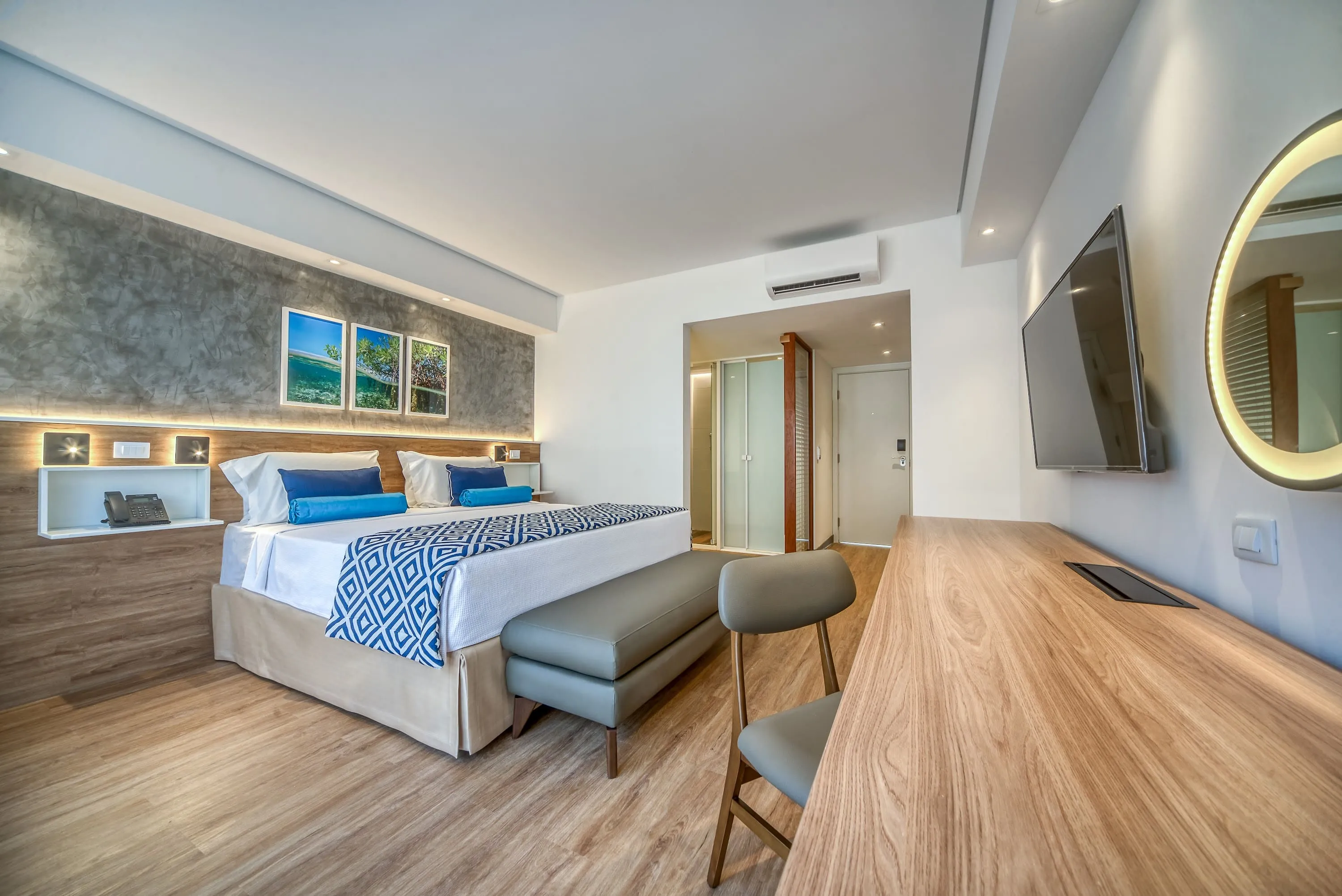 Configuración del alojamiento en el Estándar Vitória-Régia con cama queen size, mesa y silla con espejo redondo iluminado, armoniosa decoración de madera, lámparas y marcos con fotografías de las playas de Maragogi. También puede ver el baño y la entrada del apartamento.