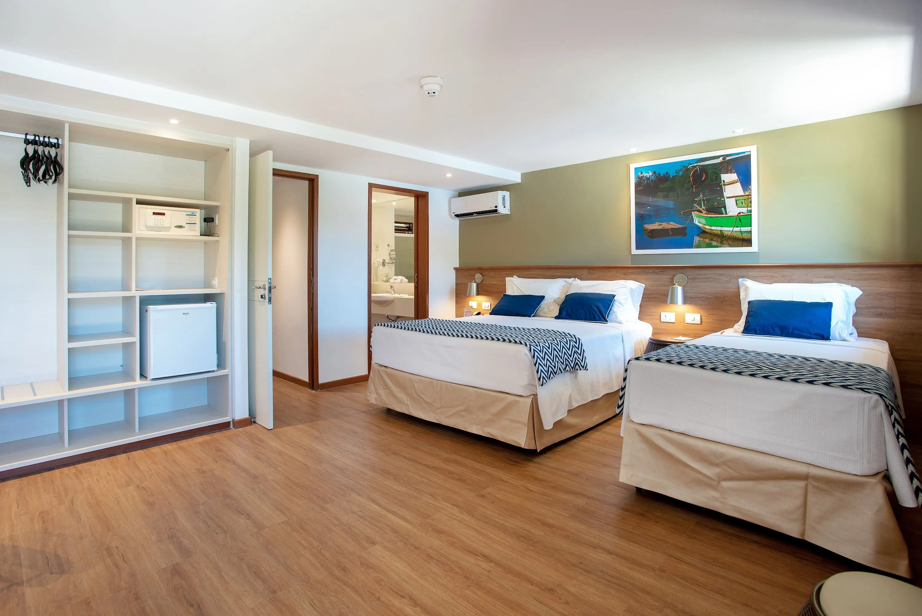 Otra configuración de la Suite Familia, alojamiento en el Salinas Maragogi Resort Todo Incluido, con una cama queen size, una cama individual, armario, minibar, lámparas, decoración en madera, una imagen de una balsa en el río Maragogi y puerta al baño.