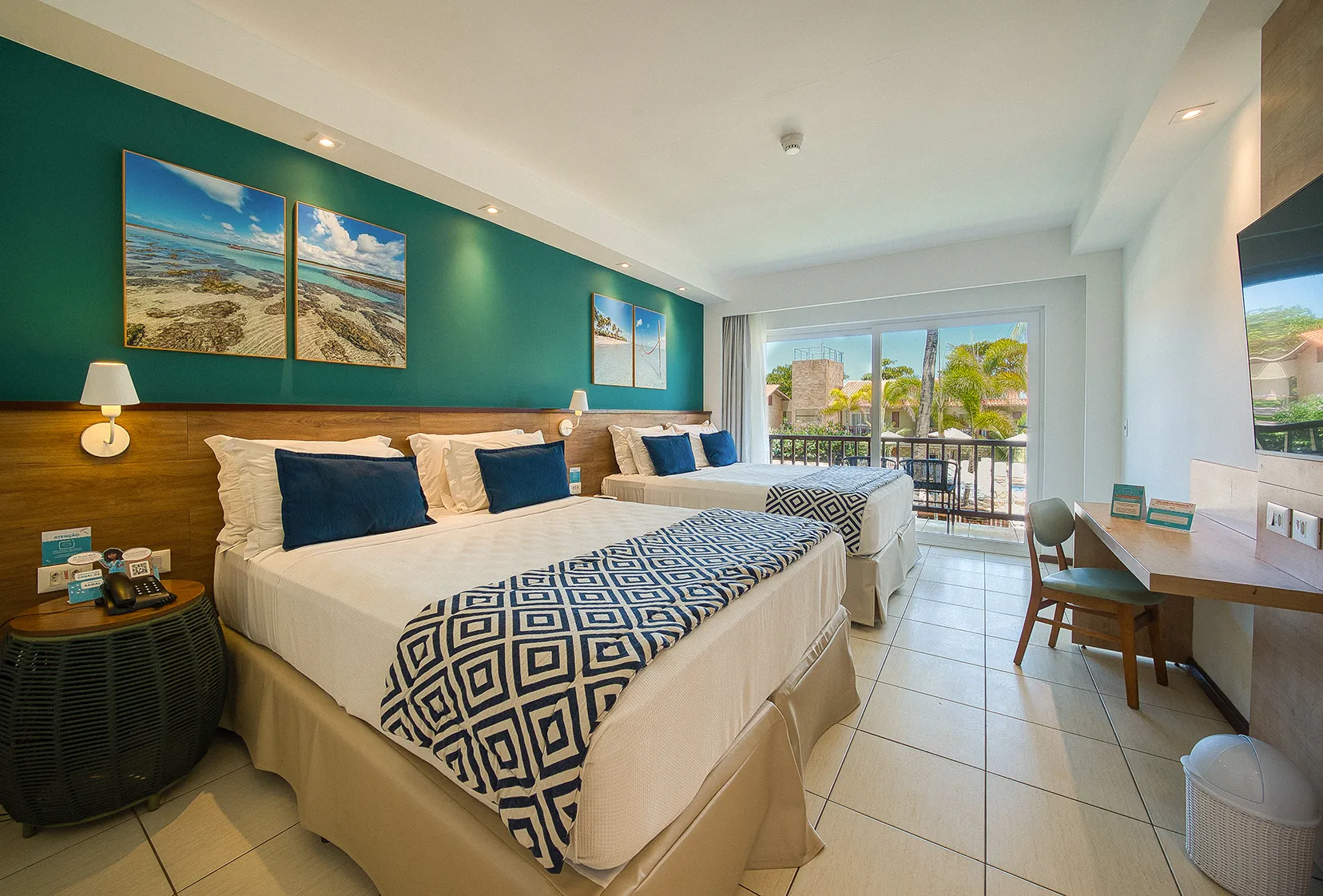 Duas camas queen size dispostas lado a lado, com duas luminárias, sendo uma para cada cama, em frente a painel de madeira com TV a cabo, mesa e cadeira.