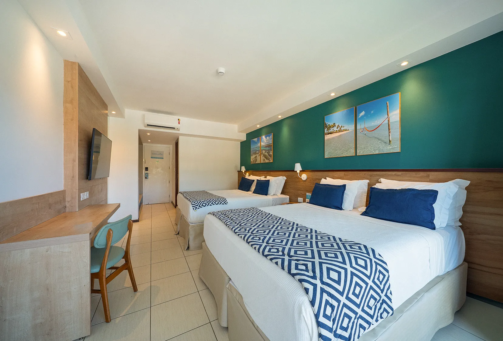 Porta de entrada da acomodação superior ao lado do roupeiro e com duas camas queen size dispostas lado a lado, duas luminárias, sendo uma para cada cama, em frente a painel de madeira com TV a cabo, mesa e cadeira.