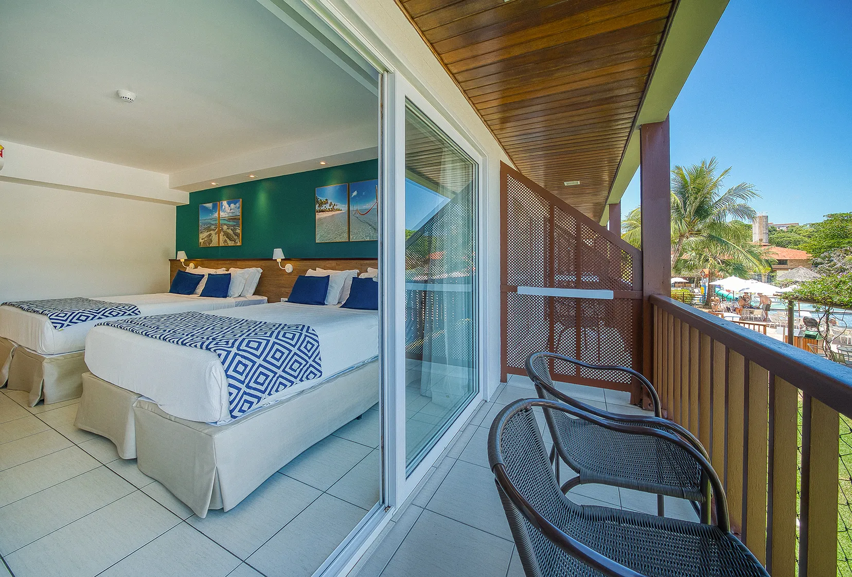 Varanda da acomodação superior, com duas cadeiras e vista para a piscina no canto. Ao fundo o quarto com duas camas king size, com parede verde-azulado e quadros pendurados.