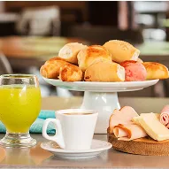 Mesa com pães, frios, queijos, café, sucos e bolo em ambiente de restaurante