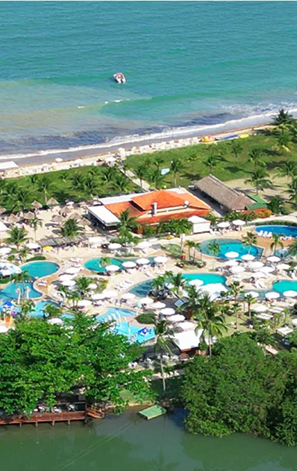 Imagem aérea do Salinas Maragogi mostrando o complexo de piscinas, dos quartos, pontos de consumo e ao fundo a imensidão da praia. Todo o resort é cercado por área verde e o um rio passa pelo meio do resort.